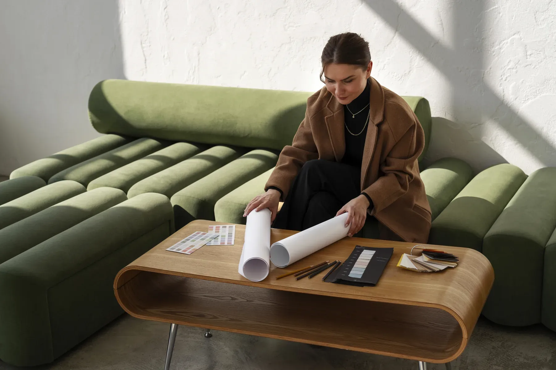 An interior designer in a camel coat unrolling floor plans on a curved wood coffee table beside fabric and finish samples