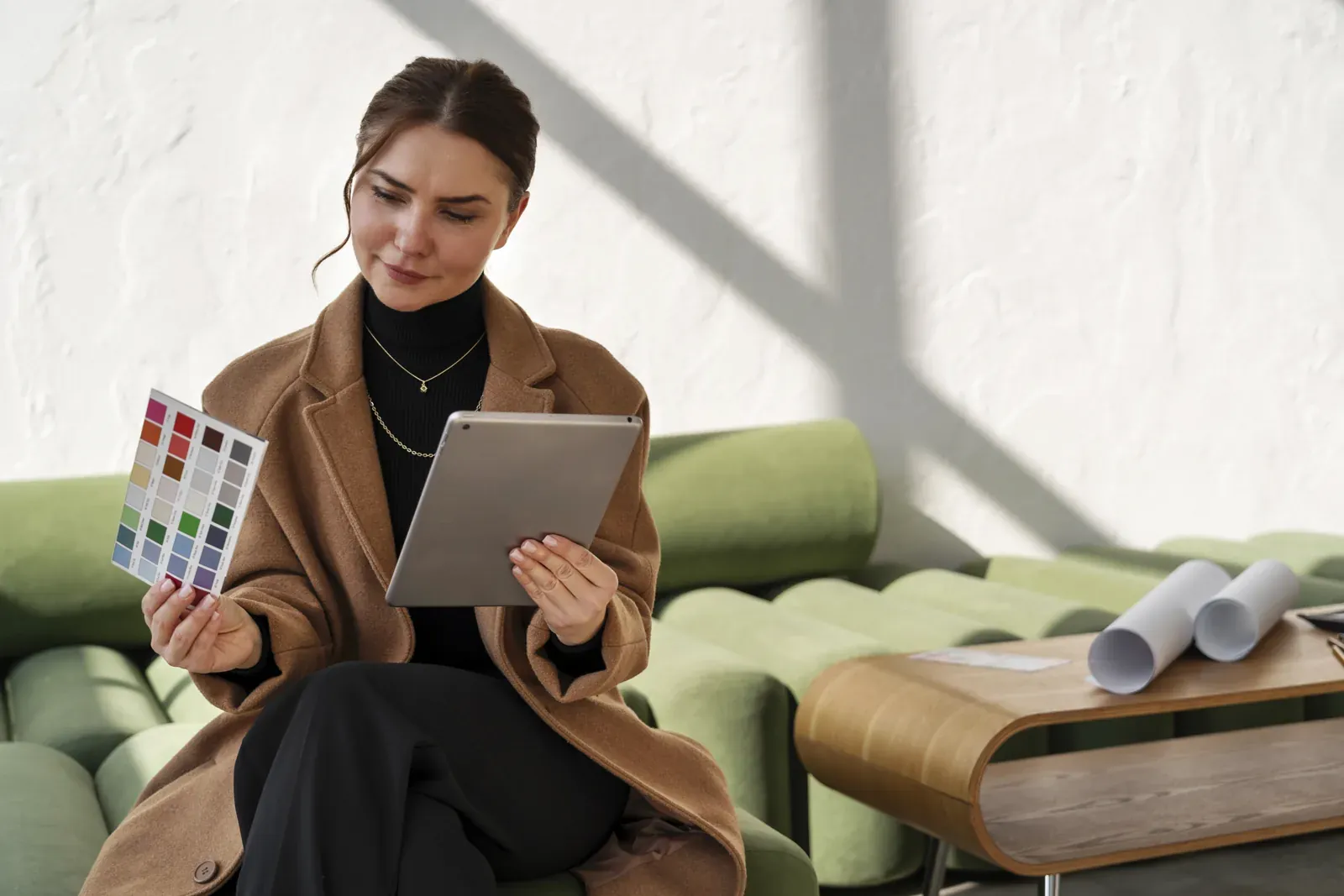 An interior designer in a camel coat frowning at her tablet while she holds up a paint color card